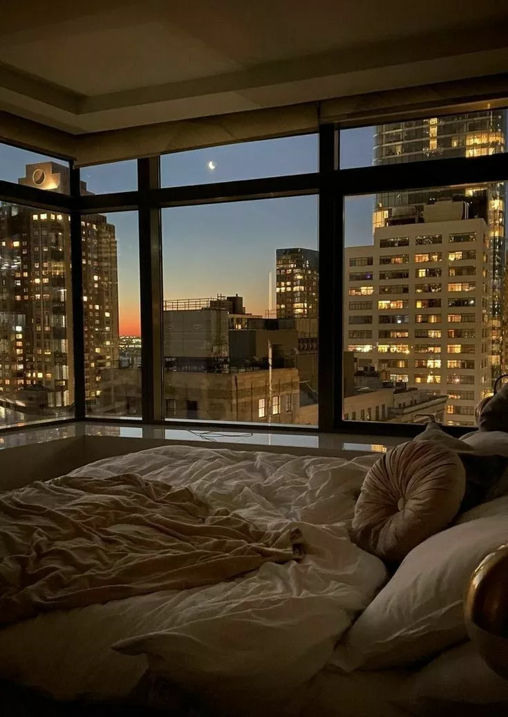 Discover the serene ambiance of a cozy bedroom, set against the breathtaking backdrop of a bustling cityscape at dusk.